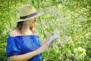 Young woman in a straw hat reading a book against the background of flowering trees in spring