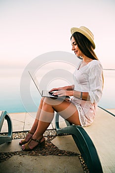 Young woman in straw hat relaxing near waterpool with laptop in the evening