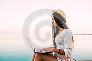 Young woman in straw hat relaxing near waterpool with laptop in the evening