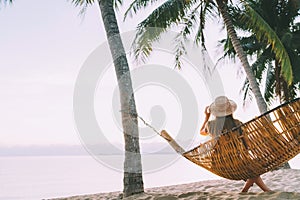 A young woman in straw hat sitting in a hammock swinging between a palm trees on the overseas island sand beach at sunrise time.