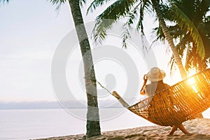 A young woman in straw hat sitting in a hammock swinging between a palm trees on the overseas island sand beach at sunrise time.