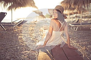 Young woman in straw hat sitting on a tropical beach, enjoying sand and sunset. Laying in the shade of palm tree parasols