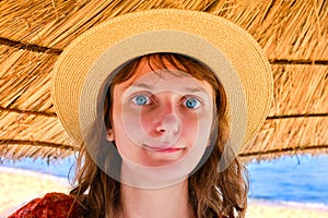 A young woman in a straw hat stands under an umbrella. Girl on the evening beach near the blue sea. Female face, close-up