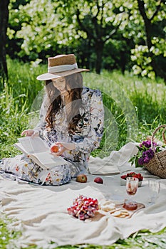 Young woman in straw hat wearing summer dress reading a book while relaxing in the park.