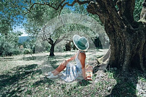 Young woman with straw sun hat under an olive tree on a background of idyllic Mediterranean landscape. Olive forest