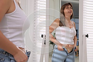 Young woman struggling to put on tight jeans near mirror at home