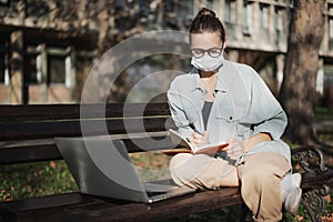 A young woman student in a facial protective mask sitting on a bench in the park and typing on her laptop on a sunny day