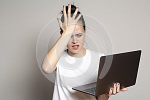Young woman student looks at her laptop, she is confused and worried about solving a problem, holds her palm on her forehead and