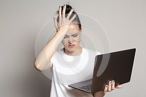 Young woman student looks at her laptop, she is confused and worried about solving a problem, holds her palm on her forehead and