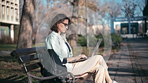 A young woman student sitting on a bench in the park and typing on her laptop