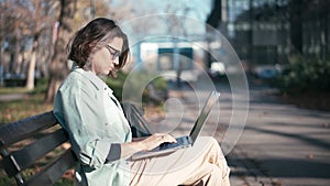 A young woman student sitting on a bench in the park and typing on her laptop