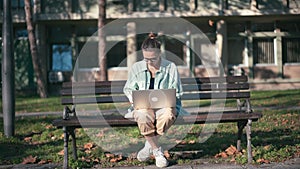 A young woman student sitting on a bench in the park and typing on her laptop