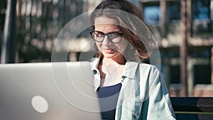 A young woman student sitting on a bench in the park and typing on her laptop