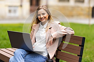 Young woman student sitting on a bench in the park and typing on her laptop on a sunny day