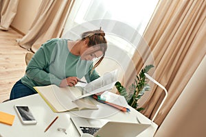 Young woman studying remotely. Young female student sitting at the table and reading handbook, using laptop, taking