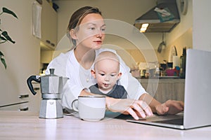 Young woman studying or working online at home while having breakfast with her baby on kitchen. Millennial mother on maternity