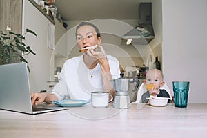 Young woman studying or working online at home while having breakfast with her baby on kitchen. Millennial mother on maternity