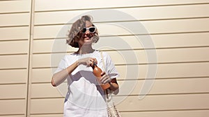 Young woman in stylish sunglasses holds string shopping bag outdoors. Smiling girl with curly hair drinks fresh water