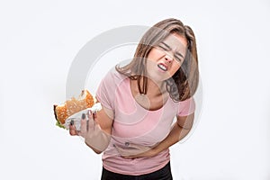 Young woman suffer from pain in stomach. She hold bitten burger. Another hand is on stomach area. Isolated on white