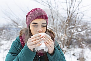 Young woman suffering from a seasonal cold and flu blowing her nose on a handkerchief as she stands outdoors on winter. Healthcare