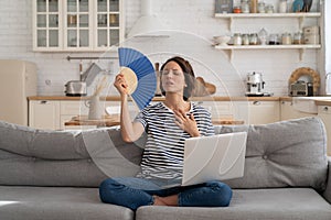 Young woman suffers from heatstroke flat without air-conditioner waving fan sitting on couch at home