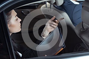 Young woman in suit sitting in backseat of car with digital tablet and talking