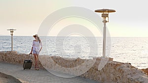 Young woman in summer clothes, sunglasses and sun hat, walks with travel suitcase along empty promenade, by the sea