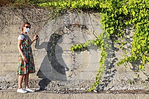 Young woman in summer dress and protective face mask alone stands next to concrete wall with ivy