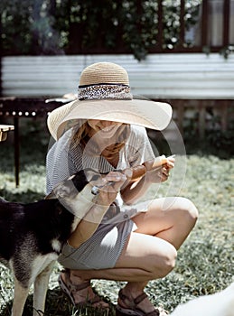 Young woman in summer hat grilling meat outdoors in the backyard, sitting with her dog