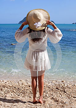 Young woman in summer white dress with straw hat looking to a sky and sea. Back view.