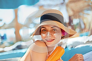 Young woman with sun cream on face holding sunscreen bottle on the beach. Female in hat applying moisturizing lotion on skin