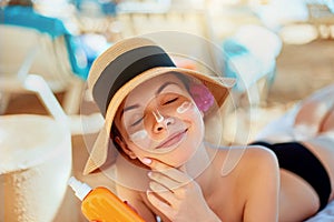 Young woman with sun cream on face holding sunscren bottle on the beach. Female in hat applying moisturizing lotion
