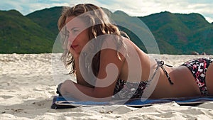 Young woman sunbathing on summer beach on green mountain background. Beautiful woman in bikini sun tanning on sandy