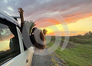 Young woman in sunglasses looks out of the window of a white car. Pretty female brunette looking to the sky