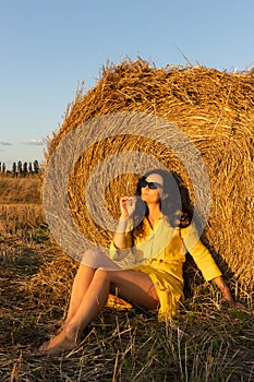 Young woman in sunglasses sitting near straw roll on field.