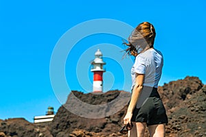 Young woman with sunglasses stand in front of a lighthouse on a rocky shore. Girl near a phare on a voulcanic rocks photo