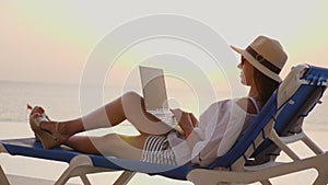 Young woman in sunglasses and sun hat, uses laptop, lying on a lounger, at the pool, at sunset or sunrise. freelancer