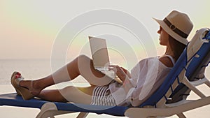 Young woman in sunglasses and sun hat, uses laptop, lying on a lounger, at the pool, at sunset or sunrise. freelancer