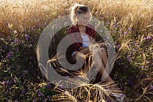 A young woman at sunset sits in a field with flowers and wheat.