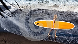 Young woman with surfboard walk on black sand beach