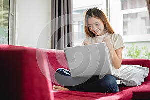 Young woman surfing the internet by laptop on red sofa and glad in successful trading in her house. Selling and online shopping