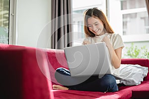 Young woman surfing the internet by laptop on red sofa and glad in successful trading in her house. Selling and online shopping photo