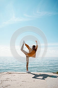 Young woman in a sweater on a beach practicing yoga Dancer`s Pose Natarajasana