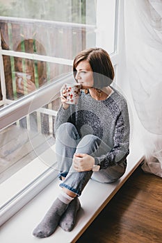 Young woman in a sweater and boyfriend jeans relaxing near big window