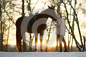 Young woman in sweater and cap standing near brown horse on snow at sunset. Background of winter trees and sunset sky