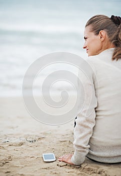 Young woman in sweater with cell phone sitting on lonely beach