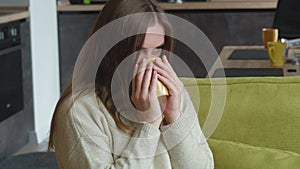 Young woman in sweater enjoys delicious hot tea while sitting on cozy sofa at home. Portrait of beautiful pensive female