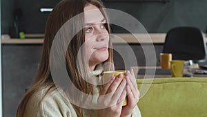 Young woman in sweater enjoys delicious hot tea while sitting on cozy sofa at home. Portrait of beautiful pensive female