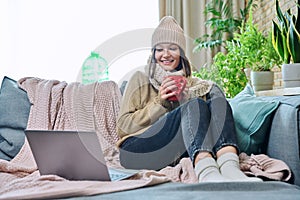 Young woman in sweater hat sitting at home, basking with cup of hot drink