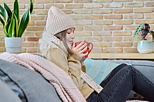 Young woman in sweater hat sitting at home, basking with cup of hot drink